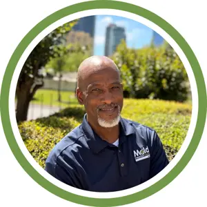 A man with a bald head and gray goatee smiles outdoors, wearing a navy blue Mold logo shirt, suggesting his expertise in mold inspection. Green bushes and city buildings shine in the background, all framed by a green and white circular border.