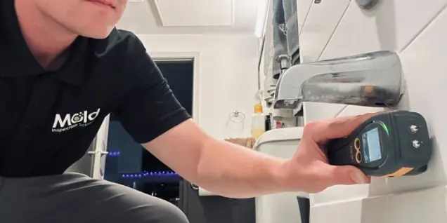 A person in a black polo shirt uses a handheld moisture meter to check tile walls near a bathroom faucet, likely conducting an inspection for mold or water damage.