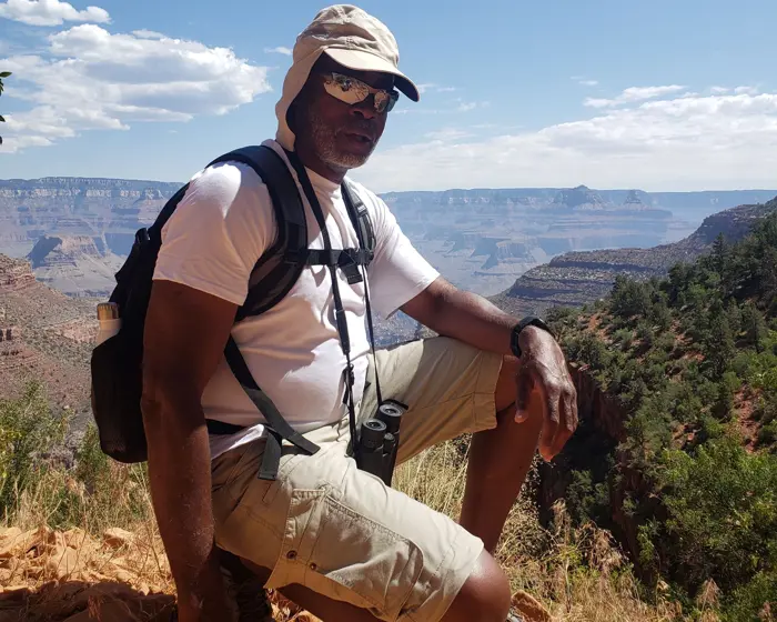 A man equipped for adventure, donning a hat, sunglasses, and backpack, poses on a rocky trail at the Grand Canyon. With an eye for detail reminiscent of inspection and testing, he marvels at the expansive canyon under scattered clouds.