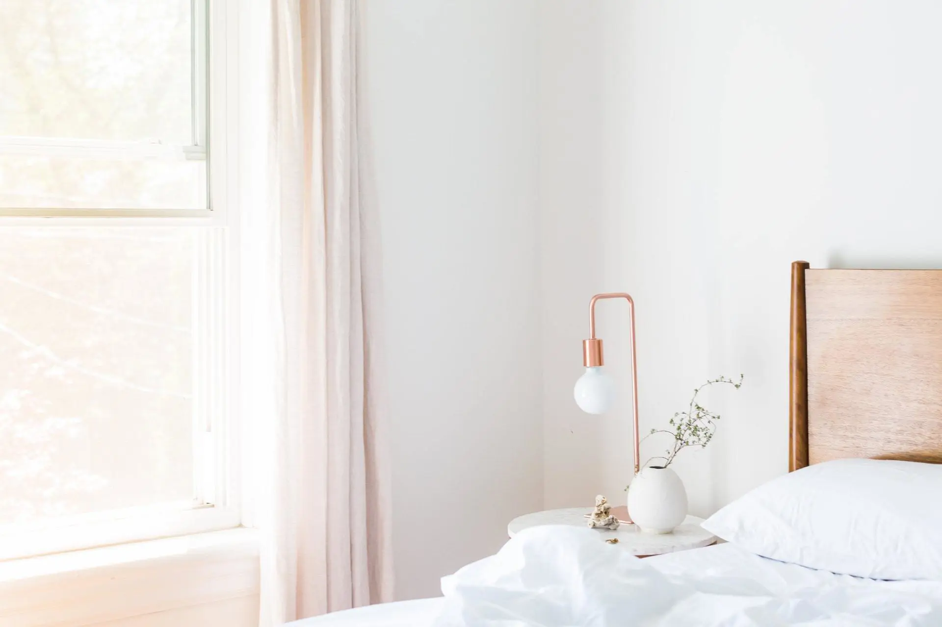 A minimalist bedroom with a wooden bed frame, white bedding, and a small round side table. The table holds a white vase with greenery and a modern copper lamp. Natural light filters through the window with sheer curtains on the left, providing an ideal setting for mold assessment in bright daylight.