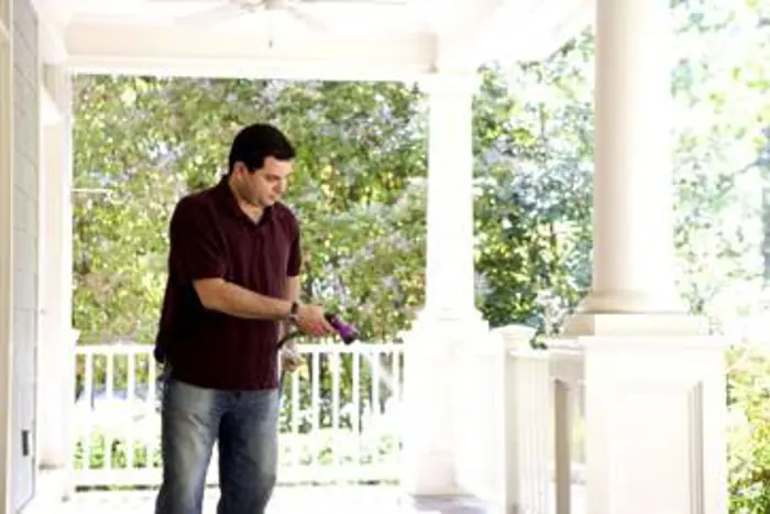 A person in a dark shirt and jeans conducts an inspection, using a cleaning spray on the railing of a sunlit porch. Large white columns and lush greenery are visible in the background, enhancing the serene atmosphere.