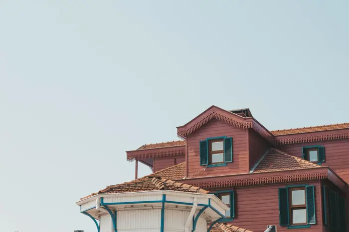 A partial view of a red building with a brown tiled roof and green shutters against a clear blue sky suggests an inspection might be due. The foreground shows the top of a white structure with blue trim, hinting at the meticulous sampling needed for mold prevention.