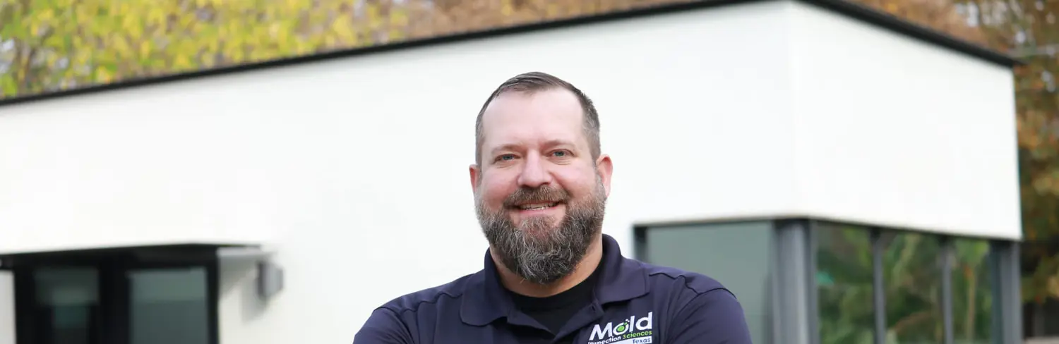 A man with a beard stands confidently with arms crossed, wearing a navy blue shirt that displays the Mold logo. In the background, a modern-style house with large windows and autumn foliage awaits an expert mold assessment to ensure its pristine condition.