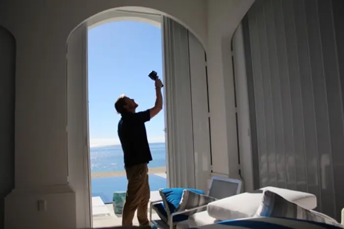A person stands silhouetted in front of a tall, open door overlooking the ocean, conducting an inspection with their camera or phone. Sunlight streams in, illuminating a modern interior with white and blue furnishings.