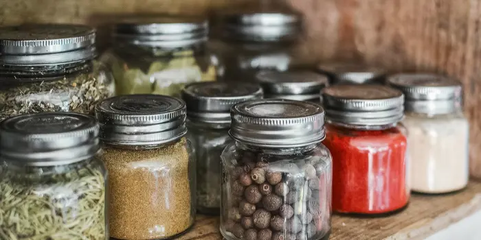 A wooden shelf holds various glass jars, perfect for sampling diverse spices and herbs. The jars have metal lids and contain colorful spices, including red pepper flakes and whole peppercorns. Natural light highlights the array of textures and colors, making it an inviting scene for any culinary assessment.