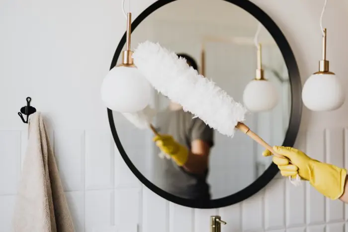 A person wearing yellow gloves carefully dusts a round mirror with a white duster in a bathroom, as if conducting an assessment of cleanliness. A beige towel hangs on a hook nearby, and two pendant lights are visible beside the mirror.