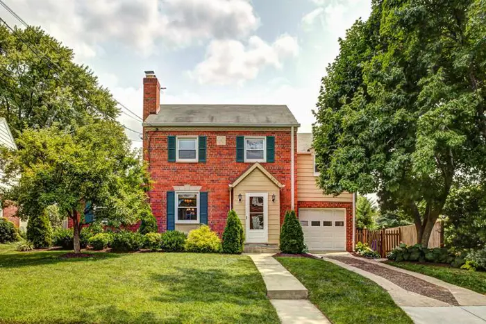 A two-story red brick house with blue shutters and a single-car garage stands proudly. Surrounded by lush green trees and bushes, its well-manicured lawn invites you in via a concrete pathway. The sky is partly cloudy, setting the scene for an idyllic location ideal for mold assessment.