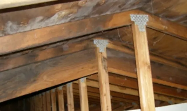 Wooden beams and trusses form the framework of a ceiling, connected with metal plates. The aged wood is observed from an angle, emphasizing the triangular connections. An investigation into its structure highlights the craftsmanship and durability of this architectural design.