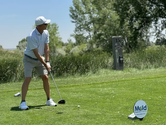 A person in a white cap and polo stands on a golf course, preparing to swing a club. The lush grassy area includes a sign for Mold Inspection Sciences. Trees and a sign marked 15 are in the background under the clear sky, framing this perfect day for both golf and mold inspection.