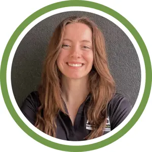 A young woman with long, wavy brown hair smiles at the camera. She is wearing a black collared shirt and is posed against a dark background, framed by a circular green and white border, reflecting professionalism in mold inspection and assessment.