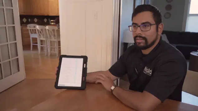 A man with glasses sits at a wooden table, holding a tablet displaying a sampling list. He wears a black collared shirt with a logo and looks toward the camera. A kitchen is visible in the background.