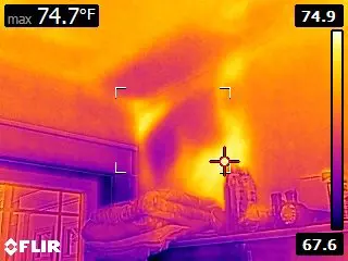 A thermal image showing a bedroom corner during an inspection. Temperature readings vary, with warmer areas in orange and cooler areas in purple, ideal for mold detection. A bed and shelves with objects are visible. Max temperature is 74.7°F.