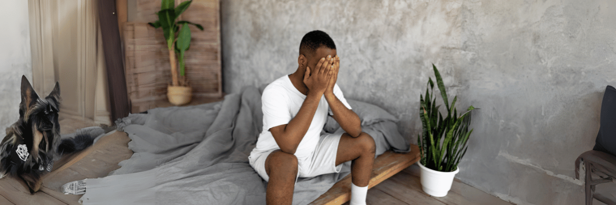 A person, clad in a white shirt and shorts, sits on the edge of a bed, covering their face with their hands as if reflecting after an intense testing day. Beside them, a dog rests on the floor. In the background, a plant in a pot and textured walls quietly watch over the scene.