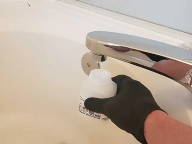 A person wearing a black glove holds a small white bottle under a running faucet in a bathroom, sampling the water. The sink and faucet are metallic and shiny, possibly part of an assessment to check for mold.