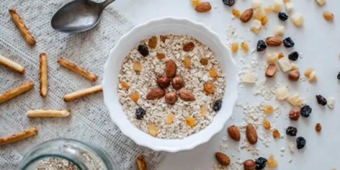 A bowl of oatmeal topped with nuts and dried fruits sits on a textured cloth, inviting a sampling of its flavors. Scattered around are nuts, raisins, and a few breadsticks. A spoon and a jar of oatmeal await nearby on the white surface.