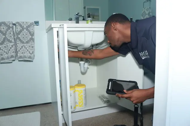 A person in a navy shirt conducts an assessment of the plumbing under a bathroom sink, holding a tablet. The cabinet door is open, revealing cleaning supplies inside. Towels hang on a nearby wall.