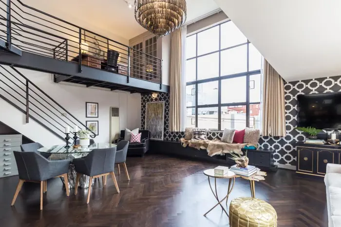A modern, stylish loft apartment with a mezzanine level awaits your inspection. It features a large window, a chandelier, dark wood floors, a round dining table with gray chairs, and a cozy seating area with pillows against geometric-patterned wallpaper.