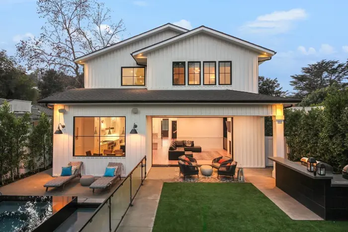 A modern two-story house with large windows, white siding, and a dark roof stands elegantly. The backyard invites inspection with its pool, green lawn, and patio area featuring lounge chairs and a fire pit. Tall trees provide a scenic backdrop.