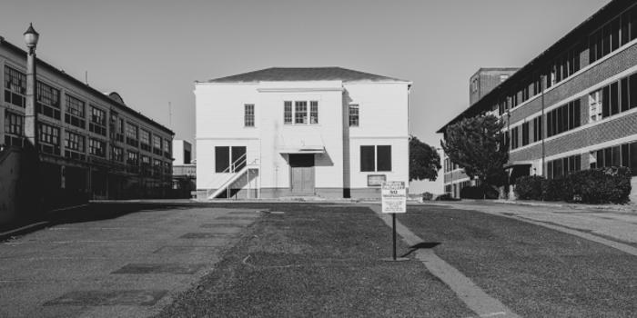 A black and white photo captures a parking lot dominated by a central white building with steps, flanked by brick structures. A parking restriction sign stands in the foreground, while the clear sky presides over this scene, evoking an air of calmness, as if time itself were under testing.