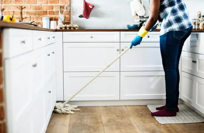 A person conducting an inspection, wearing a plaid shirt, jeans, and rubber gloves, is mopping the wooden floor in a kitchen with white cabinets and a brick wall. Various household items are visible on the countertop.