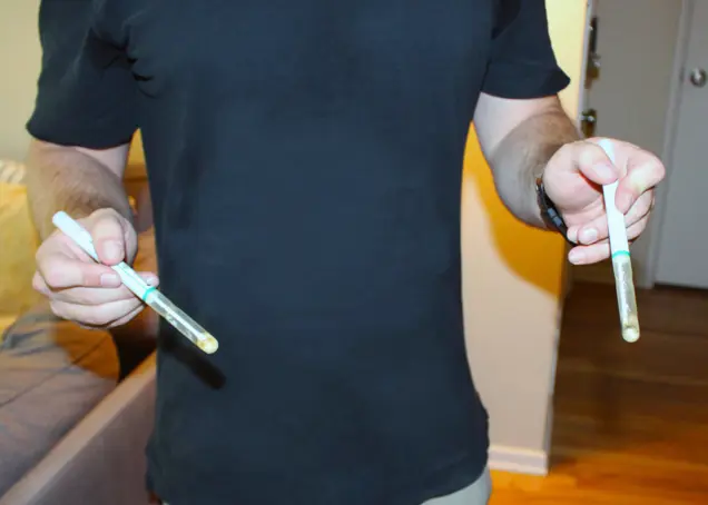 A person in a black shirt holds two test tubes, each containing a small, cloudy liquid, as they conduct mold testing indoors near a couch.