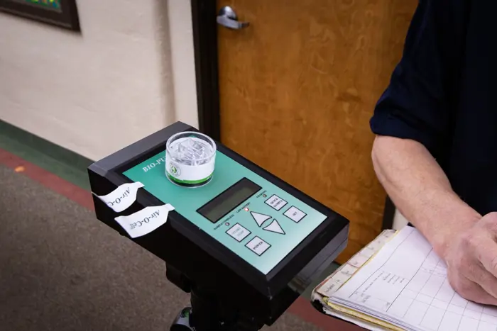 A person holding a clipboard stands beside a BIO PAK machine undergoing testing. The device features buttons and an LED screen, with a small container labeled Alcohol on top. Two additional Alcohol labels are nearby, indicating thorough assessment processes.