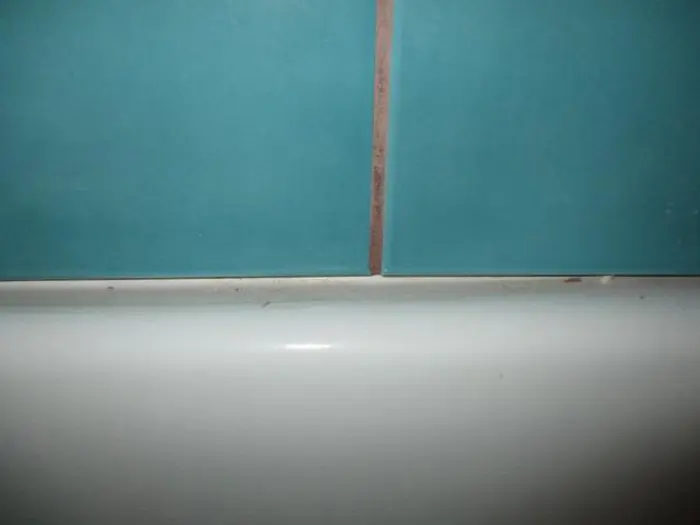 Close-up of a turquoise tiled wall with a narrow beige grout line. Subtle signs of mold can be seen along the edges. The bottom part reveals a smooth white surface, possibly a bathtub or countertop, displaying slight wear or discoloration along the edges.
