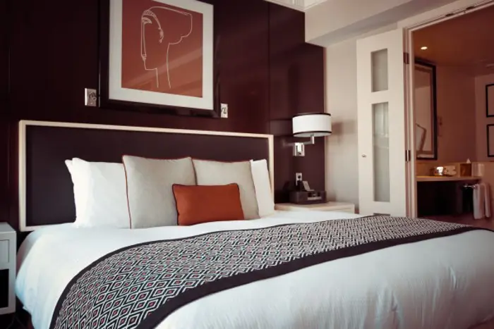 A modern hotel room featuring a neatly made bed with white pillows and blankets, accented by a patterned bed runner and an orange cushion. The room has dark wooden walls, a framed abstract art piece, and a glimpse of an adjoining bathroom ready for your investigation.