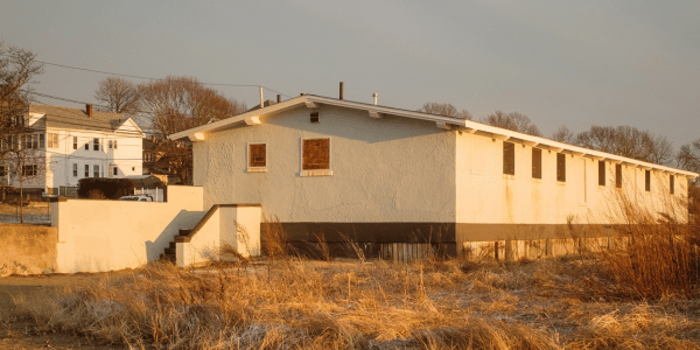 A small, flat-roofed concrete building with boarded windows sits in a grassy area, possibly awaiting mold testing. Next to it are some residential houses in the background under a clear sky on a sunny day.