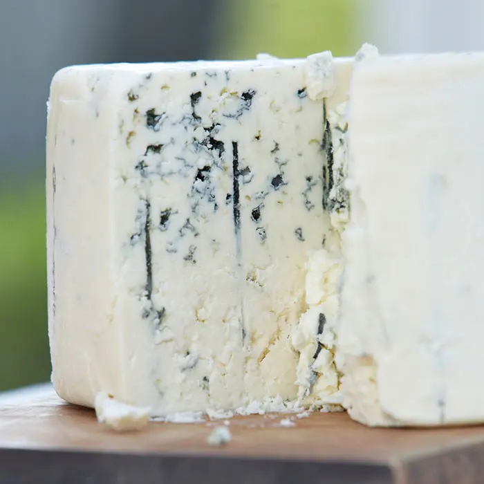 Close-up of a wedge of blue cheese on a wooden surface, showcasing its creamy white texture with mold-like blue veins running through it. The background is softly blurred, highlighting the cheese.