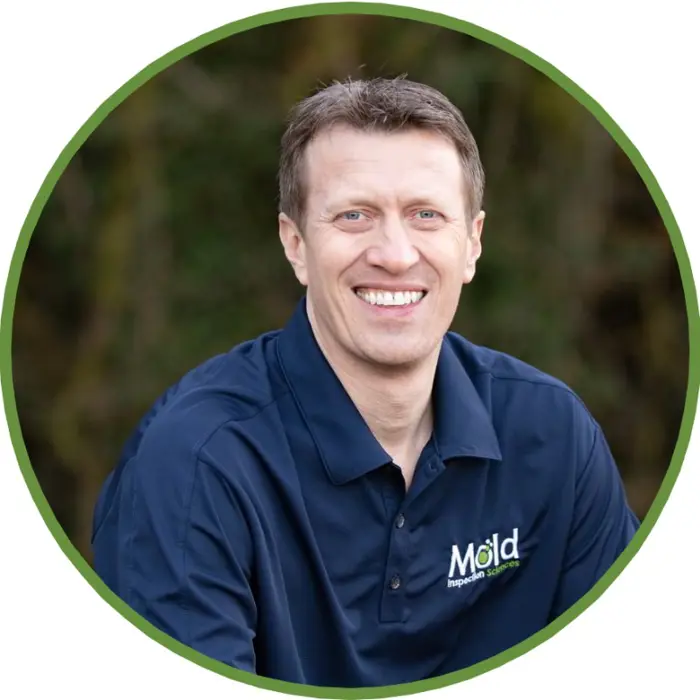 A man in a navy blue polo shirt with Mold Inspection Sciences embroidered on it smiles at the camera, embodying professionalism. Framed in a circular green outline, the image focuses on him while the blurred greenery background hints at thorough mold inspection expertise.