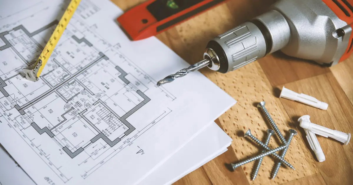 A close-up view of architectural blueprints on a wooden table, accompanied by a measuring tape, red level tool, and power drill. Nearby are screws, wall anchors, and a drill bit, indicating an assessment for a construction or renovation project.