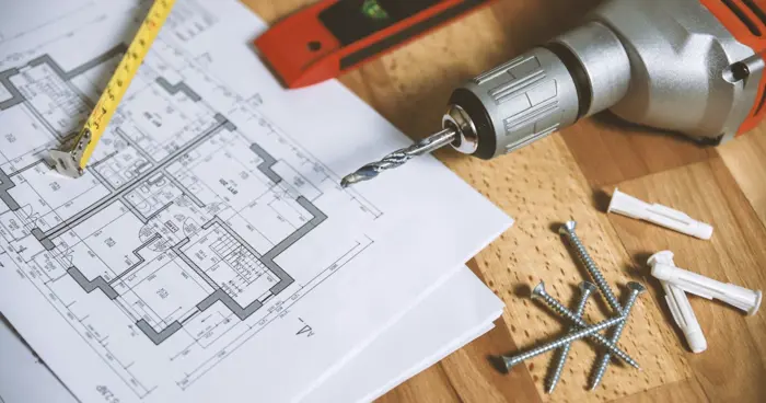 A close-up view of architectural blueprints on a wooden table, accompanied by a measuring tape, red level tool, and power drill. Nearby are screws, wall anchors, and a drill bit, indicating an assessment for a construction or renovation project.