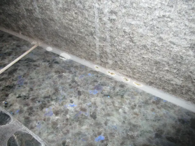A close-up view of a corner where a gray stone tile wall meets a speckled countertop, with a bead of white sealant showing small yellowish spots along the seam, possibly indicating mold.