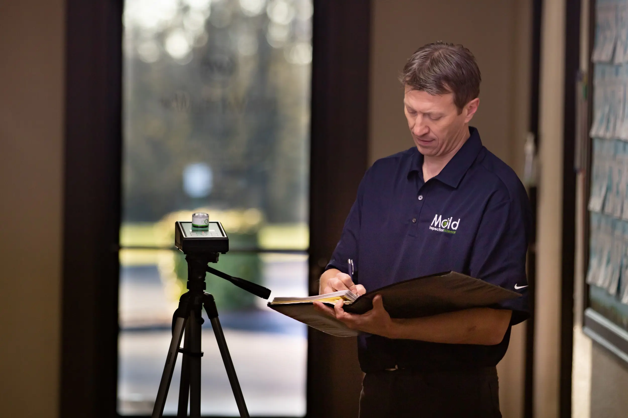 A man in a navy blue polo shirt stands indoors, conducting an inspection and writing on a clipboard. There's a tripod-mounted device nearby, with sunlight streaming through a glass door in the background.