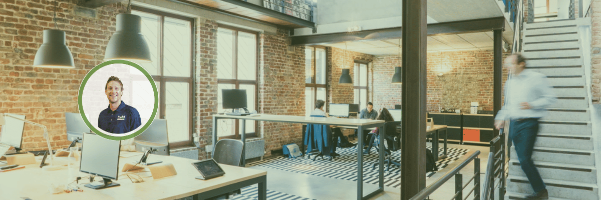 Modern office space with brick walls, wooden floors, and large windows. Several desks with computers facilitate testing new ideas, a staircase on the right, and people working. A man in a blue shirt is pictured in an inset circle on the left.