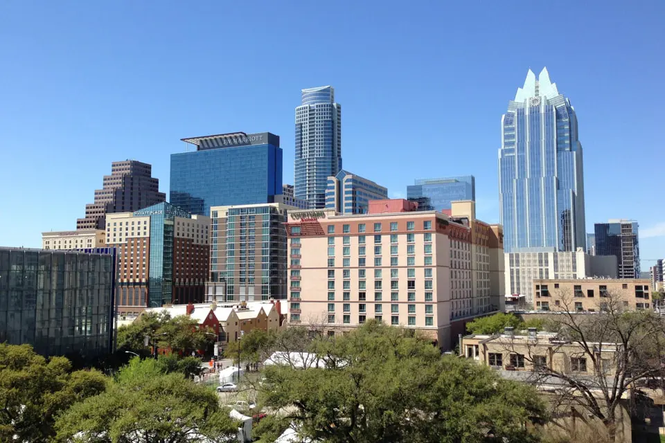 A vibrant city skyline features tall modern skyscrapers and lightly scattered clouds in a clear blue sky. An inspection of the scene reveals trees and low-rise buildings in the foreground, creating a dynamic urban landscape.