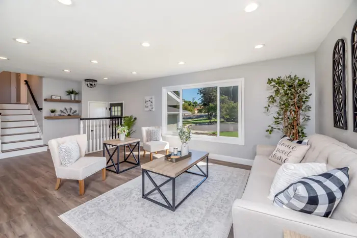 A bright, modern living room awaits your inspection, showcasing a large window, a beige sofa with decorative pillows, two armchairs, and a wooden coffee table amidst lush plants. The light wooden flooring complements the gray rug perfectly, and a staircase is visible in the background.