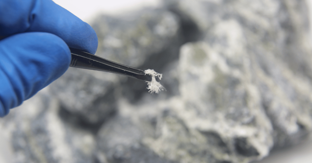 Close-up of a gloved hand holding tweezers, delicately sampling a small, fibrous white material from a blurred, rocky background.