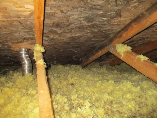 An attic space with exposed wooden beams and yellow insulation covering the floor is ready for an assessment. A silver ventilation duct extends through the insulation, and the ceiling features wooden boards.