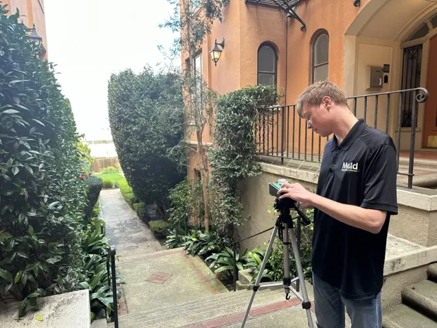 A man in a black Meld shirt adjusts a camera on a tripod beside the steps of an orange building, conducting an assessment with plants and a paved walkway visible in the background.