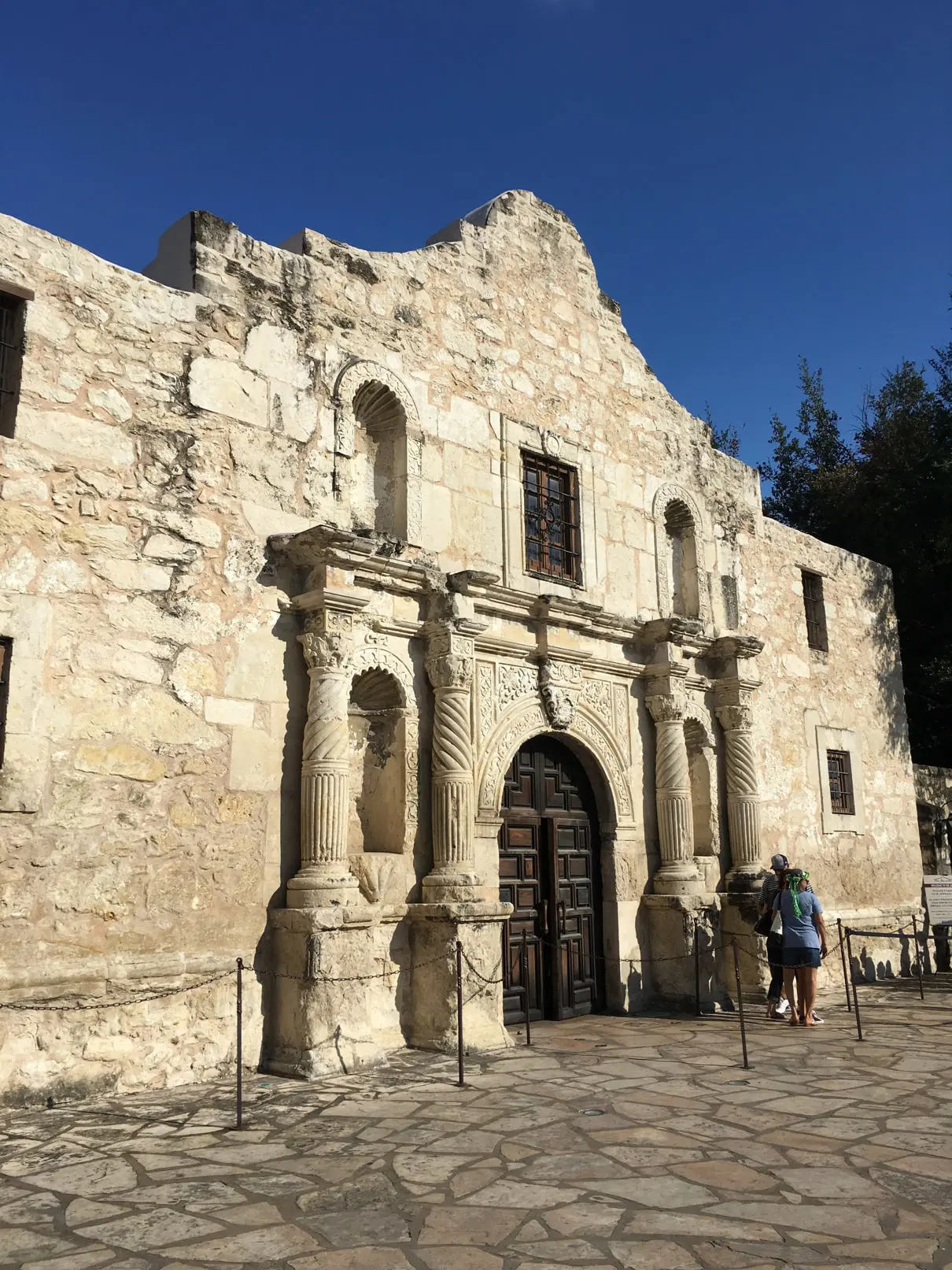 The image showcases the Alamo Mission in San Antonio, a historic Spanish mission with a rustic stone facade, arched doorway, and iconic columns. Visitors stand outside under a clear blue sky, assessing its timeless beauty.