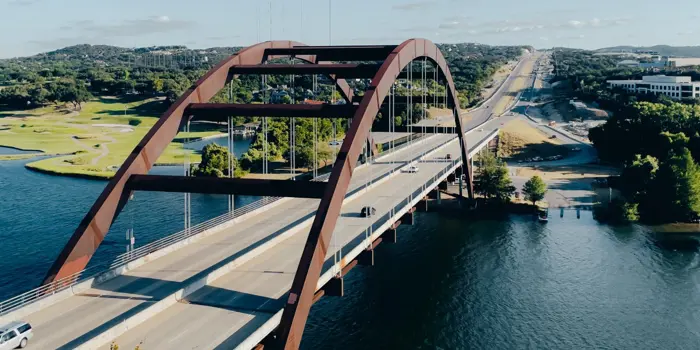 An aerial view of a large arch bridge spanning over a body of water reveals its robust design. The bridge, important for thorough inspection and testing, supports a roadway with cars traveling in both directions. It is surrounded by lush green trees and distant hills under a partly cloudy sky.