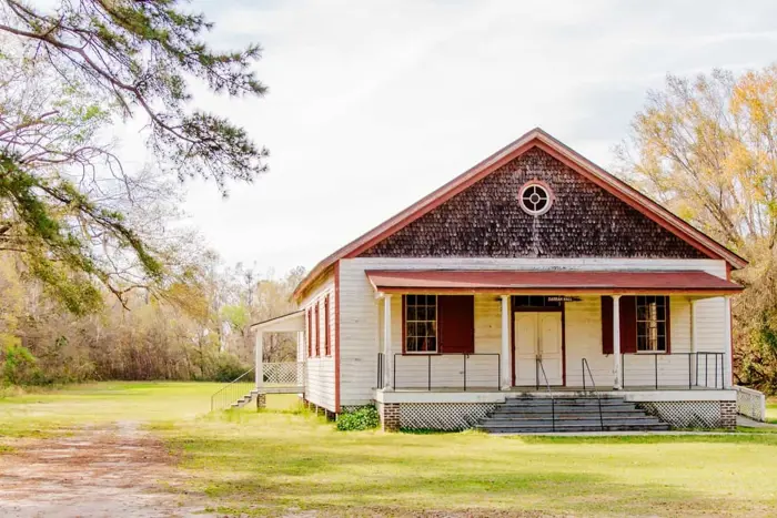 A rustic, single-story wooden building with red shutters and a symmetrical front staircase, set in a grassy field, stands as if molded by time itself. The backdrop includes trees and a cloudy sky. A porch wraps around the side, adding to its quaint charm.
