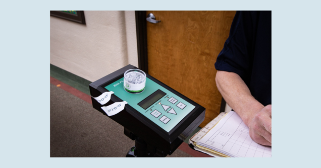 A person is standing beside a portable measuring device with a small cup on top, conducting an assessment. The device has a green display with buttons and labeled paper notes. The person is holding a clipboard with papers, next to a wooden door.