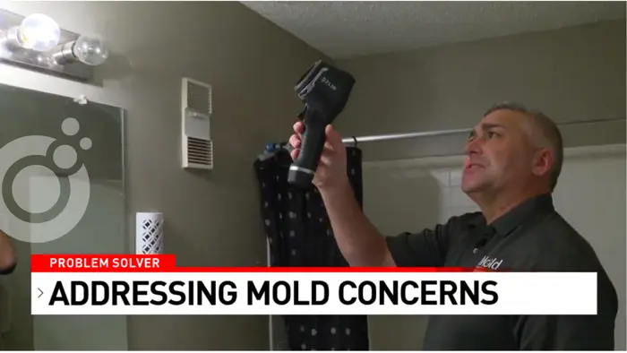 A man conducts a mold investigation with a thermal imaging camera in the bathroom, inspecting potential concerns. The space features a vent, vanity lights, and a shower curtain. Text on the image reads: Problem Solver: Addressing Mold Concerns.
