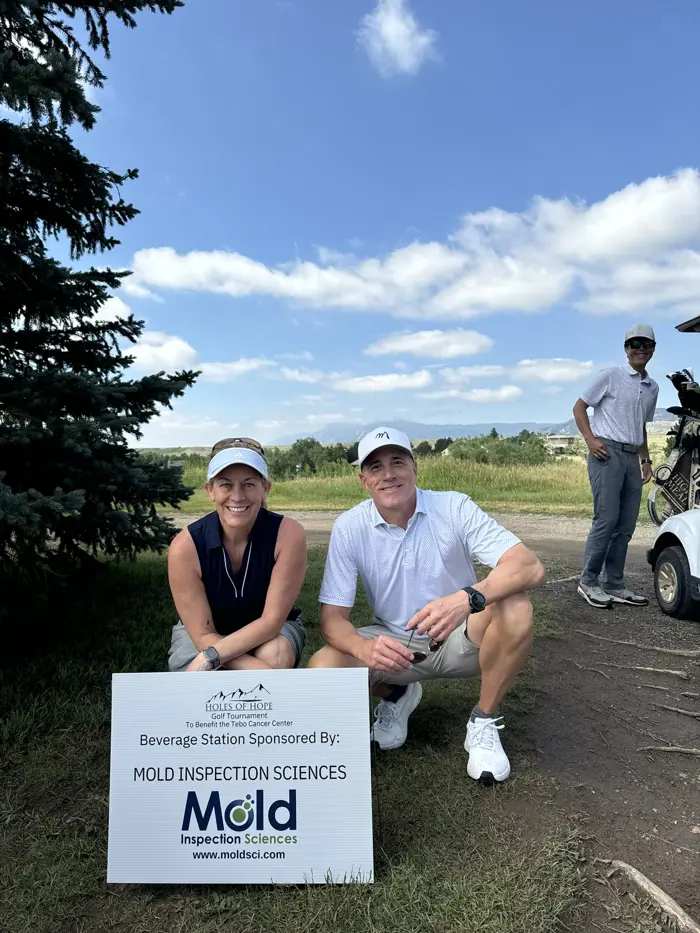 Two people kneel beside a sign on the golf course reading, "Beverage Station Sponsored By: Mold Inspection Sciences." A man stands nearby with a golf cart in the background. It's a sunny day with blue skies—perfect for mold-free assessments!