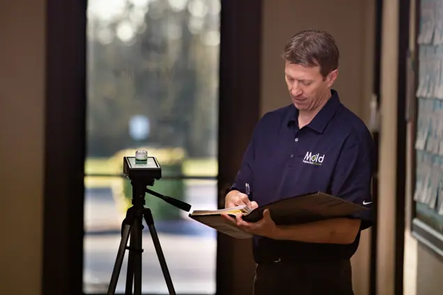 A man wearing a black polo shirt with "Mold" emblazoned on it stands indoors, conducting an assessment with his clipboard. Nearby, a tripod holds a sampling device. Light streams through the window behind him, illuminating his focused work.