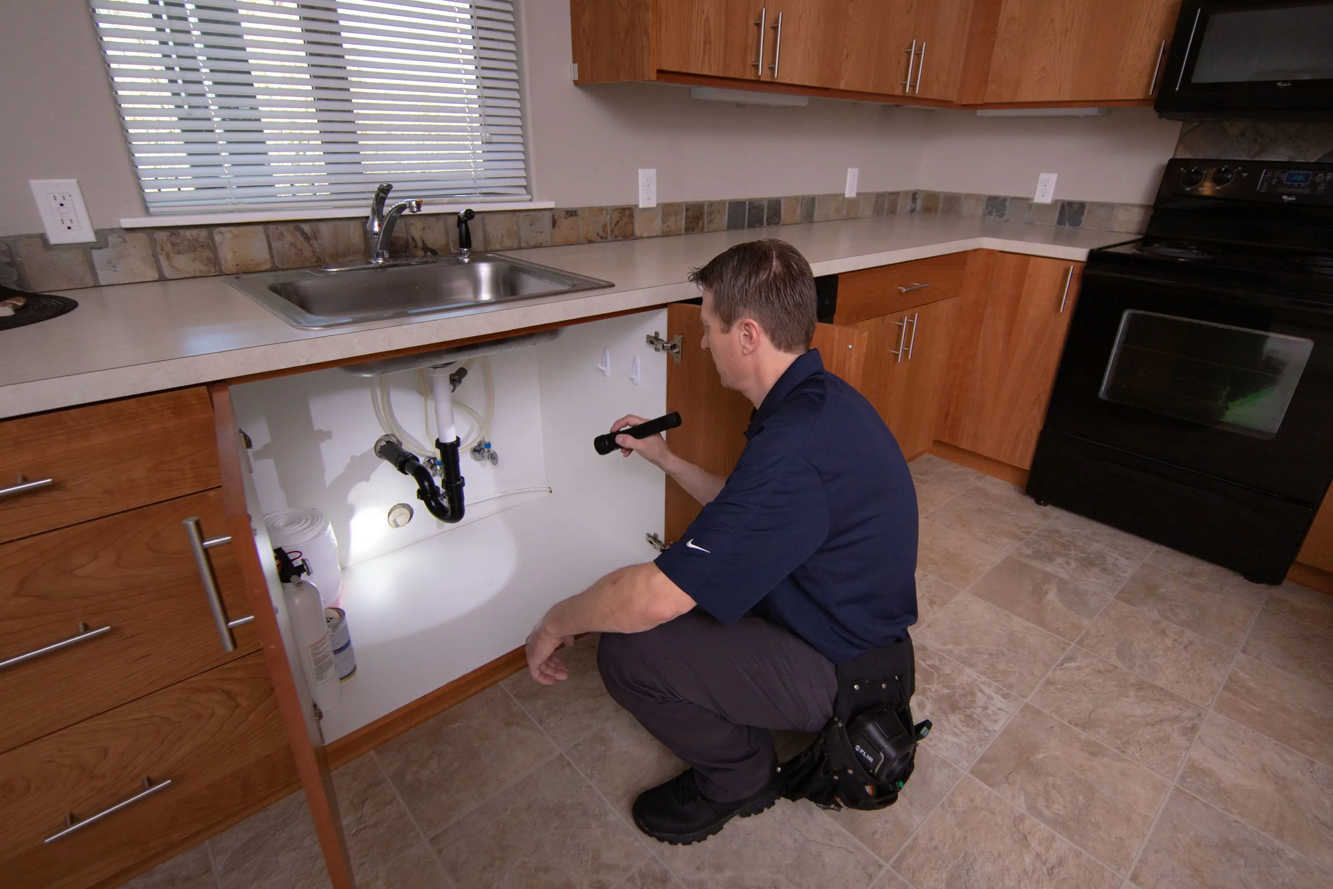 A man in a navy blue shirt and dark pants kneels on the kitchen floor, using a flashlight for a thorough inspection of the pipes under the sink. The well-maintained kitchen features wooden cabinets and a tiled floor, setting the scene for his careful assessment.