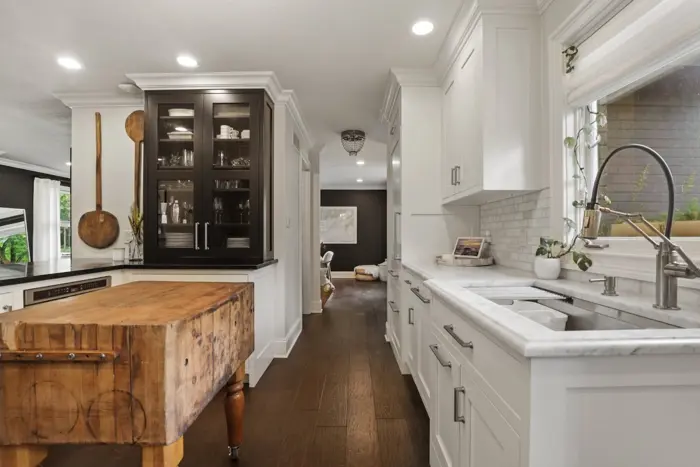 A modern kitchen with white cabinets, marble countertops, and a farmhouse sink. A wooden island stands in the center, complemented by glass-fronted cabinets on the left. Recently, an assessment revealed no mold issues. A hallway leads to a dining area in the background, lit by recessed lighting above.